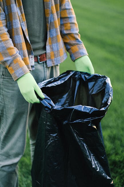 A group of large plastic bags filled with rubbish, positioned on the edge of a sidewalk in front of a black metal fence with vertical bars. The bags appear to contain mixed waste materials, possibly c